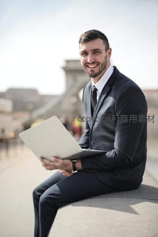 西装男坐桥边微笑持电脑