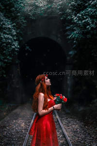红裙女子手持玫瑰，伫立于绿意盎然的隧道铁轨上仰望天空。