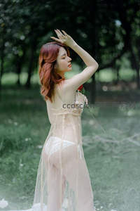 红发女子穿薄纱裙举手遮阳立林中