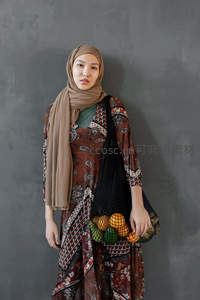 头巾女子手提网袋水果静立灰墙前