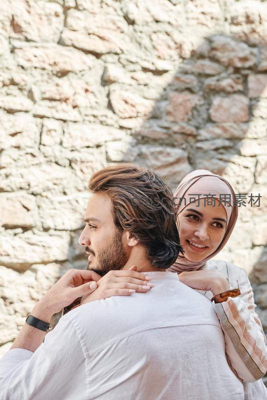 石墙前的温柔依偎，女子轻抚爱人肩头笑容灿烂