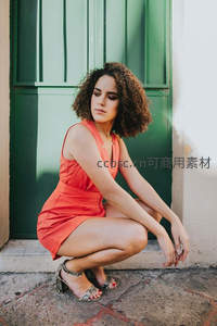 卷发女子蹲坐绿门前，橙裙优雅
