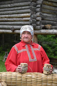 传统服饰老妇人持木杯，木屋前展现民俗风情
