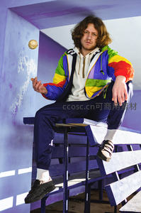 彩虹夹克男坐紫凳，玩球姿态俏皮搞怪