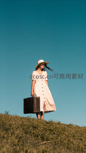 白衣女子手提旧箱伫立青草坡顶