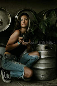 女子手持相机蹲坐酒桶旁绿植间