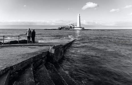 黑白影像中情侣远眺孤岛灯塔，海浪轻拍堤岸