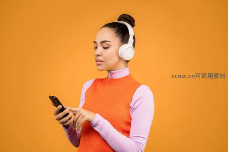 橙色背景下的时尚女性，佩戴耳机专注操作手机