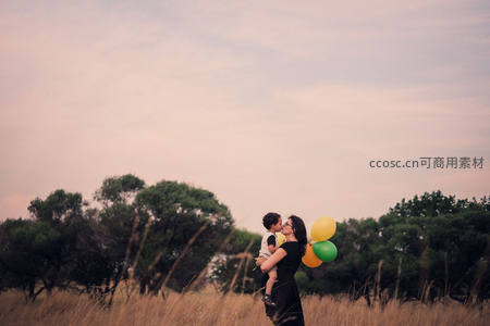 母亲怀抱婴儿亲吻，手握彩色气球于田野间