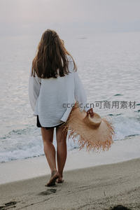女子海边漫步，手持草帽背影悠然