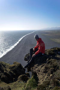 黑沙滩畔，红衣旅人静坐悬崖眺望无垠大海