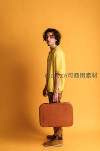 黄色背景前，卷发青年手提复古行李箱回眸