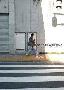 行人过马路低头看手机，阳光洒在水泥墙边