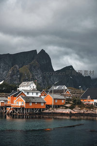 橙白渔屋临水而建，背景黑岩峻岭压境，阴云低垂渲染北欧渔村苍劲氛围
