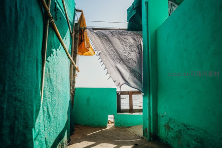 青绿色棚屋巷道中，挂毯随风轻扬，透见海面波光