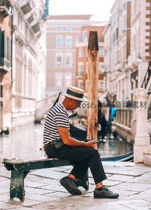 威尼斯水巷边戴草帽男子静坐长椅，专注看手机