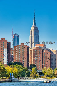 纽约帝国大厦矗立蓝天之下，红砖公寓与绿树环绕水岸
