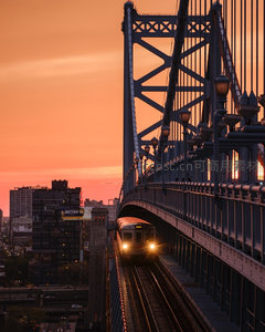 橙红暮色中列车穿行钢桁架大桥，车灯如星点亮工业诗意夜幕