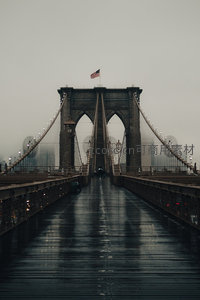 雨雾中的布鲁克林大桥，湿漉漉的桥面倒映着哥特式塔楼与飘扬的星条旗