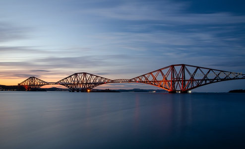 福斯桥钢铁巨构剪影于暮色水面上，橙红灯光点亮百年铁路奇迹