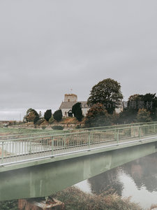 雾气弥漫河畔，绿色人行桥横跨水面，远处古教堂静立树丛间