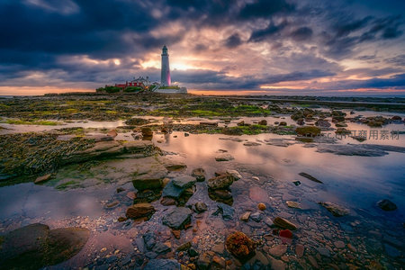 日落时分灯塔矗立礁石海岸，云霞绚烂倒影水中
