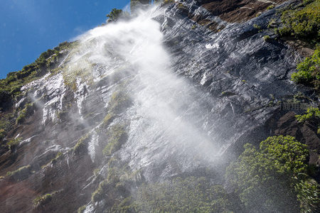 阳光穿透水雾的悬崖瀑布，飞溅水珠在蓝天下闪耀光芒