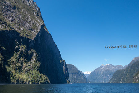 开阔峡湾远景，双峰对峙间雪顶远山浮现，碧水如镜