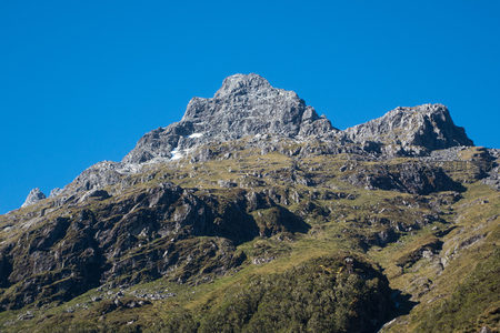 巍峨山峰耸立蓝天之下，岩壁陡峭，草甸点缀其间
