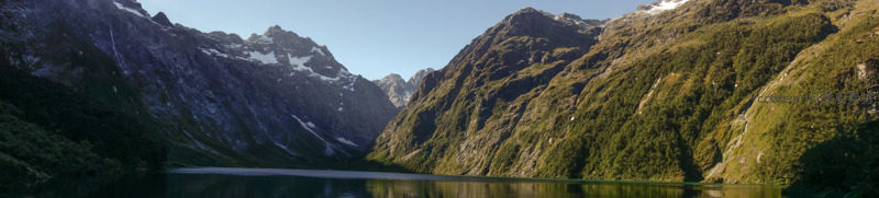  panoramic view of alpine lake flanked by forested cliffs and snow-dusted peaks under clear blue sky