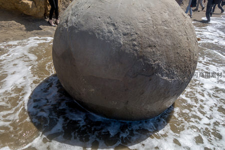 潮水漫过摩拉基巨石底部，游客脚边浪花飞溅显生动瞬间