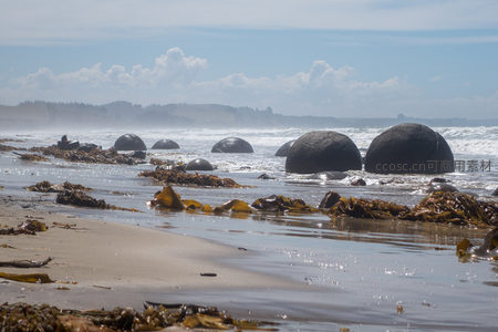 远景视角下的莫拉基巨石阵，沙岸延伸至林线山丘，云影浮动