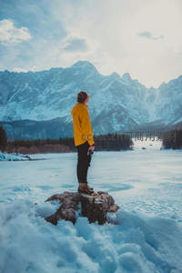 黄衣旅人伫立冰湖岩礁，仰望雄伟雪山与冬日静谧天际
