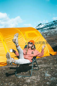 橙帽女子躺椅上举杯欢笑，身后亮黄帐篷映衬蓝天雪岭