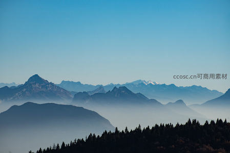 层峦叠嶂的阿尔卑斯山脉在晨雾中若隐若现，近处松林墨绿，天空澄澈湛蓝