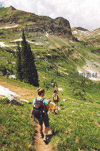 徒步者携犬穿行绿意盎然的高山草甸，雪迹斑驳，松林点缀其间
