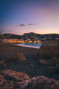 暮色海岸小镇灯火初上，黑沙滩与巨岩映衬紫霞余晖