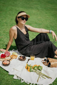 黑裙女子戴遮阳帽静坐草坪，椰碗水果与向日葵环绕，简约优雅野餐风