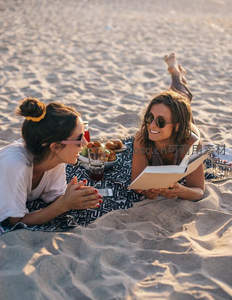 两位女性沙滩野餐共读闲聊，夕阳下放松享受假日宁静时光