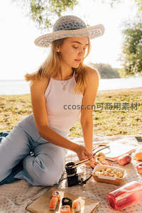 金发女子湖畔野餐专注摆盘寿司，草帽相机与便携餐盒点缀自然