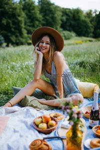 金发女子戴宽檐帽微笑望向镜头，野餐场景温馨愉悦