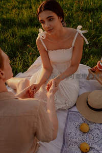 情侣草地对坐牵手：白衣女子凝视伴侣，野餐布与草帽映衬温柔时刻