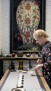 金发女子专注茶艺演示，长桌列白瓷杯，背景为大型花卉漆画屏风