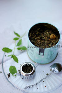 薄荷绿茶叶罐开盖特写，散落干叶与金属茶滤置于玻璃盘上，清新自然