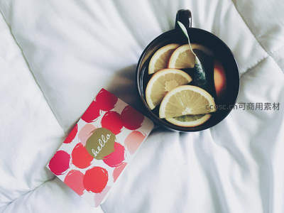 黑瓷杯中柠檬片与茶包浸润，旁置波点贺卡于洁白被褥之上