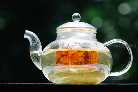 特写透明玻璃茶壶内金盏花舒展于清亮茶汤中，背景虚化绿意盎然