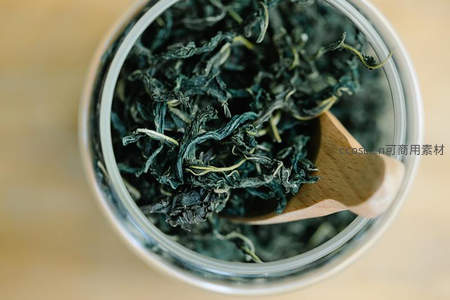 玻璃罐中深绿卷曲干茶叶特写，木勺轻舀，展现传统茶叶储存与质感