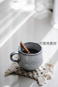 灰釉粗陶杯盛热茶，肉桂枝斜倚杯沿，窗影洒落编织杯垫