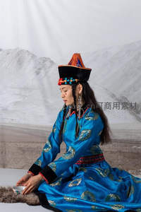 蒙古族女子身着蓝金刺绣盛装，头戴彩珠高帽，静坐雪山背景前品茶