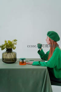 红发女子戴绿贝雷帽，侧坐桌前啜饮茶，旁置水培绿植与玻璃茶壶